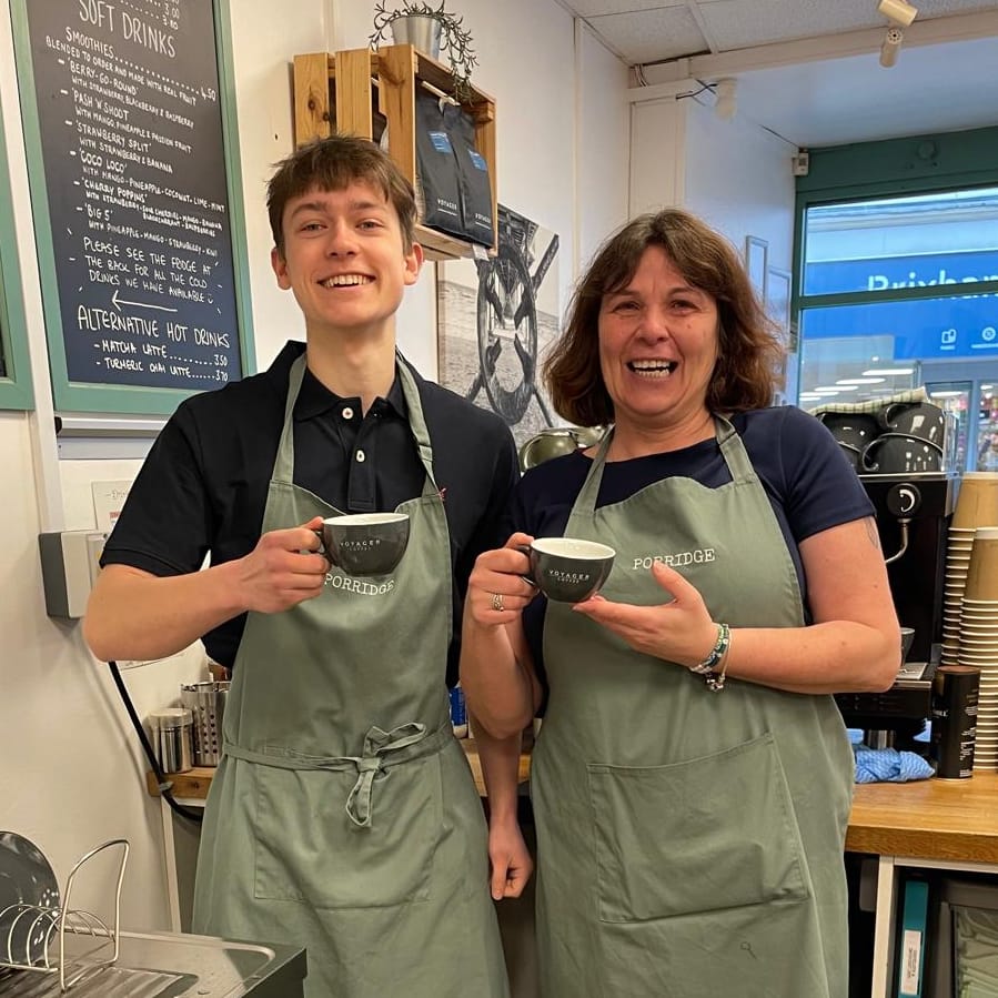 Clare and Cal at the Porridge counter in PORRIDGE-embroidered sage aprons, holding Voyager Coffee cups in front of the smoothie chalkboard