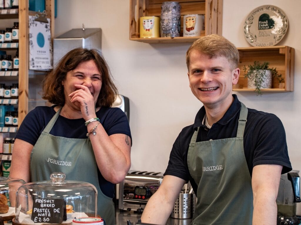 Clare and Cal behind the counter at Porridge, laughing, with a freshly baked Pastel de Nata under a glass cake dome on the bar in front of them